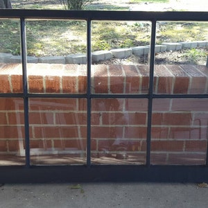 May include: A rectangular, black-framed window with eight panes of clear glass. The window is resting against a red brick wall. The window is approximately 48 inches wide and 24 inches tall.