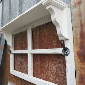 May include: White painted wooden shelf with a window pane design and two black metal hooks. The shelf is mounted on a wall with a rusty metal surface.