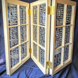 May include: A wooden framed seating chart, resembling a window, with multiple panes. Each pane displays names written in white, organized by table number. The chart is hinged and stands upright, ideal for weddings or events.