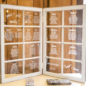 May include: A wedding seating chart displayed on a set of repurposed windows. The windows are painted white and have a rustic wood frame. The seating chart is written in white calligraphy and lists the names of guests for each table number. The text at the bottom of the chart reads "#INDIANATOOSKANNA 11.11.2017".