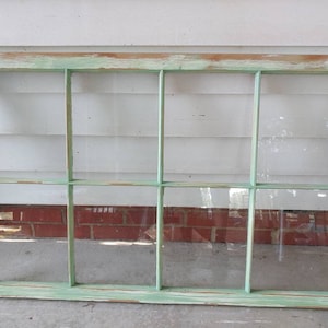 May include: An antique window frame with eight panes. The wooden frame is painted in a distressed, faded green, with visible wood grain and a weathered appearance. The glass panes are clear, and the window is resting on a concrete surface.