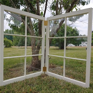 May include: A pair of white painted, wooden window panes with glass inserts. The panes are hinged together and are open to reveal a view of a grassy field and trees.