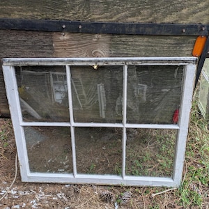 May include: A rectangular, six-pane window with a white painted wooden frame and wire mesh. The window is resting on a bed of dry grass and pine needles. The background features weathered wood.