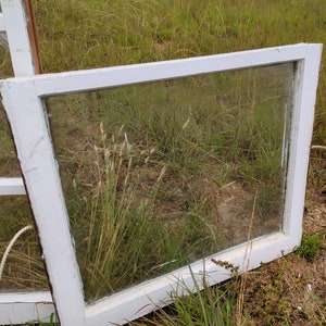 May include: A white painted wooden window frame with a single pane of glass. The window is lying on the ground in a grassy area.