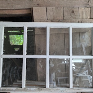 May include: A white, wooden window frame with nine panes of glass. The window is old and weathered, with some of the paint chipped away. The window is set against a backdrop of wooden planks.