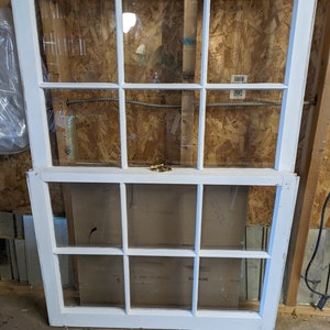 May include: Two white painted wooden windows with multiple panes of glass. The top window is closed and the bottom window is open. The windows are in a rustic setting.