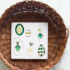 May include: A white ceramic tile with six embossed egg designs, each with a unique green and yellow pattern. The tile is set inside a woven wicker basket, creating a rustic and artistic display.