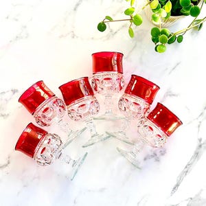 May include: A collection of eight vintage wine glasses with ruby red rims and clear, patterned bowls. The glasses are arranged on a white marble surface, showcasing their elegant design. The stems are clear and delicate, adding to the overall sophistication.
