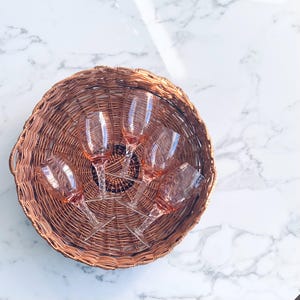May include: A woven basket holds five pink-tinted wine glasses. The glasses have clear stems and bowls, arranged in a circular pattern. The basket is made of brown wicker and sits on a white marble surface.