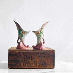 May include: A pair of vintage ceramic vases with a unique, asymmetrical shape. The vases feature a light pink base that transitions to a cream color, with a flared top. Green leaves and red berries decorate the front. The vases are displayed on a wooden crate.