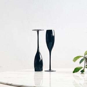 May include: Two black champagne flutes on a white surface. One flute has a wide, flat base and a tall, slender stem. The other has a traditional flute shape. The flutes are reflective and sit in front of a white background.
