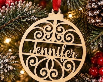 May include: A circular light-colored wooden Christmas ornament featuring intricate laser-cut scrollwork patterns and the name Jennifer written in a cursive script across the center, suspended by a thin red ribbon.
