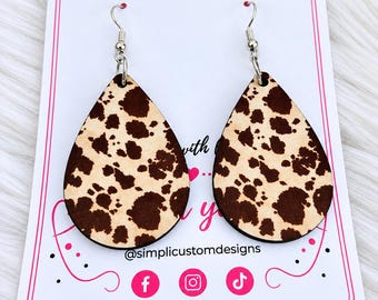 May include: A pair of teardrop-shaped earrings featuring a brown and cream cow print pattern. The earrings are attached to a white display card with the text @simplicustomdesigns and social media icons for Facebook, Instagram, and TikTok.