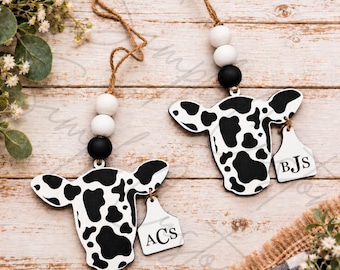 May include: Two cow-shaped wooden ornaments featuring a black and white spotted pattern. Each ornament hangs from a jute twine string with two white beads and one black bead. A small white ear tag is attached to each, displaying the letters ACS or BJS.
