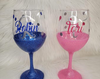 May include: Two clear wine glasses with glitter-coated stems and bases. One glass features blue glitter with the name Robin in blue script and a silver monogram. The other features pink glitter with the name Tori in pink script and a silver monogram.