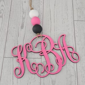 May include: A pink wooden monogram with the letters KSA, hanging from a jute string. Above the monogram are three decorative wooden beads in white, pink, and black. The background is a light gray wooden surface.