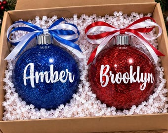 May include: Two round Christmas ornaments in a cardboard box. One blue glitter ornament features the white text Amber and a blue and white ribbon. The other red glitter ornament features the white text Brooklyn and a red and white ribbon.
