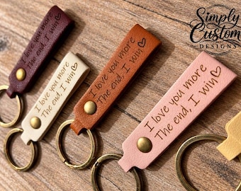 May include: Four rectangular leather keychains in dark brown, cream, tan, and light pink, each featuring a brass-toned metal rivet and a circular keyring. The text "I love you more The end, I win" with a heart symbol is engraved on each.