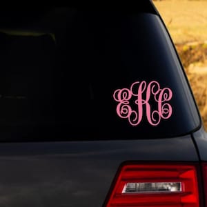 May include: A light pink vinyl decal featuring a decorative, interlocking monogram of the letters E, K, and C in a cursive script font, applied to the dark glass surface of a vehicle rear window above the red taillight assembly.