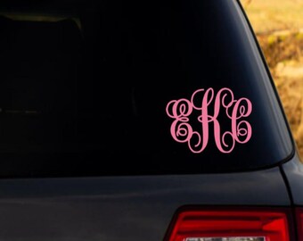 May include: A light pink vinyl decal featuring a decorative, interlocking monogram of the letters E, K, and C in a cursive script font, applied to the dark glass surface of a vehicle rear window above the red taillight assembly.