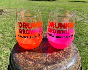 May include: Two stemless wine glasses with a gradient finish, one in orange and one in pink. Both glasses feature the text "DRUNKIN GROWNUP AMERICA RUNS ON WINE" printed in bold red and pink lettering on the front surface of the clear glass.