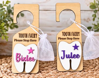 May include: Two light-colored wooden door hangers with a cutout hook. Each features the text "TOOTH FAIRY Please Stop Here" above a white tooth-shaped cutout. One has a pink star and the name "Bixlee" in pink, the other a purple star and "Julee" in purple. Each includes a small white organza drawstring pouch.