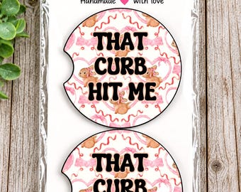 May include: Two circular car coasters in clear plastic packaging. The coasters feature a repeating pattern of pink hearts, ribbons, and brown bunnies. The black text on each coaster reads, THAT CURB HIT ME. The header card says Car Coasters.