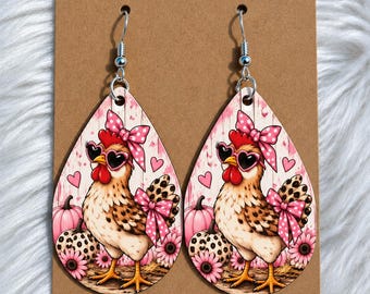 May include: Teardrop-shaped earrings featuring a graphic print of a chicken wearing heart-shaped sunglasses and a pink polka-dot bow. The design includes pink pumpkins, leopard print accents, and small pink hearts on a light wood-toned background.