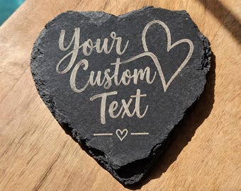 May include: A heart-shaped black slate coaster with rough, uneven edges. The surface is engraved with the text "Your Custom Text" in a cursive font, accompanied by a large outline of a heart on the right and a small heart centered below the text.