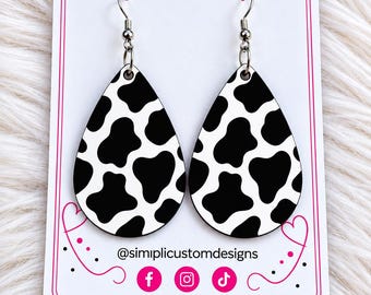 May include: A pair of teardrop-shaped earrings featuring a black and white cow print pattern. The earrings are displayed on a white card with the text @simplicustomdesigns and icons for Facebook, Instagram, and TikTok printed at the bottom.