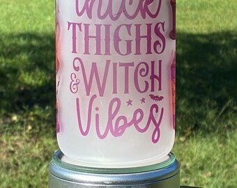 May include: A frosted glass can-shaped tumbler with a bamboo lid, featuring the purple text "Thick THIGHS & WITCH Vibes" along with a witch hat and bat graphic. The tumbler is displayed on a silver circular stand.