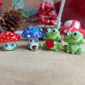 Könnte beinhalten: Vier kleine Glasfiguren, zwei grüne Frösche, ein roter und weißer Pilz und ein blauer und weißer Pilz. Die Frösche haben große Augen und ein Frosch hält ein rotes Herz.