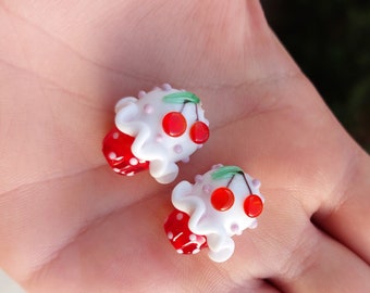 May include: Two white glass cupcake charms with red and green cherries. The cupcakes have white frosting and red polka dots.