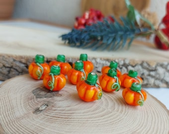 Può includere: Una collezione di dieci piccole perline di vetro a forma di zucca con steli verdi e vernice arancione e bianca. Le perline sono disposte su una superficie di legno.