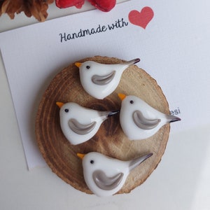 Peut inclure: Quatre oiseaux en verre blanc avec des ailes grises et des becs orange sont disposés sur une tranche de bois ronde. Les oiseaux sont faits à la main et la tranche de bois porte le texte "Handmade with" et un cœur rouge.