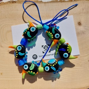 May include: A colorful glass bead necklace with black, green, blue, and yellow accents. The beads are shaped like cartoon faces with large eyes and a variety of expressions. The necklace is strung on a blue cord.