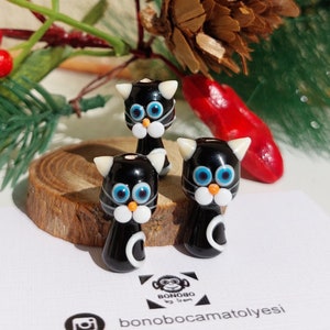 Puede incluir: Tres figuritas de gato de vidrio negro y blanco con ojos azules. Los gatos están sentados sobre una superficie de madera.