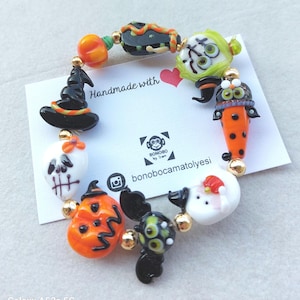 Op de afbeelding: Een Halloween-armband met kleurrijke glazen kralen. De armband bevat kralen in de vorm van een heksenhoed, pompoen, schedel en andere feestelijke ontwerpen. De kralen zijn geregen met goudkleurige accenten.