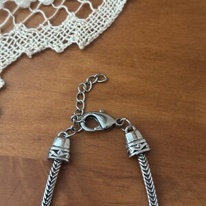 May include: A silver-toned necklace with a herringbone chain design. The necklace features decorative end caps and a lobster clasp closure. The background includes a wooden surface and a white lace doily.