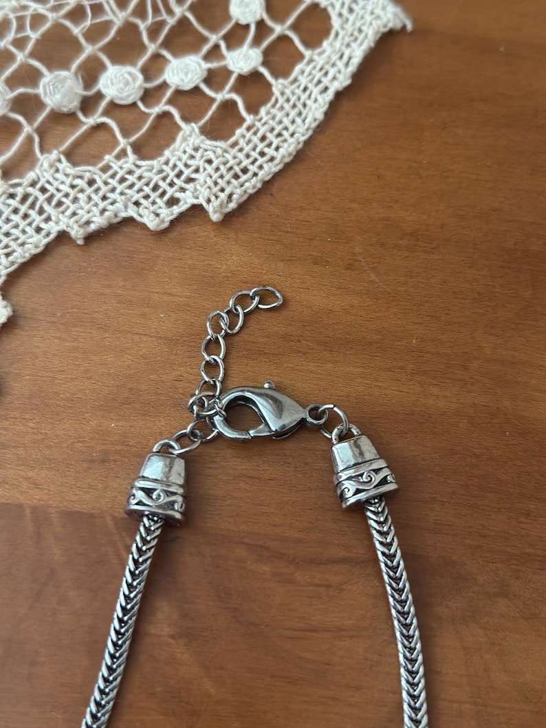 May include: A silver-toned necklace with a herringbone chain design. The necklace features decorative end caps and a lobster clasp closure. The background includes a wooden surface and a white lace doily.
