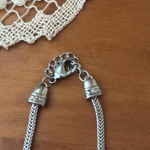 May include: Close-up of a silver necklace with a herringbone chain design. The necklace has a lobster clasp and decorative end caps. A white lace doily is in the background, resting on a wooden surface.