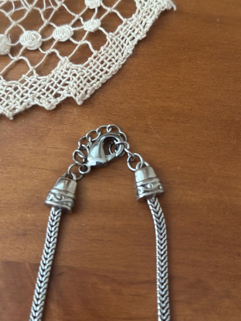 May include: Close-up of a silver necklace with a herringbone chain design. The necklace has a lobster clasp and decorative end caps. A white lace doily is in the background, resting on a wooden surface.