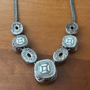 May include: Silver-toned necklace featuring a series of decorative elements. The necklace has a chain-like design with circular and square pendants. The pendants have intricate patterns and a central design. The necklace is displayed on a wooden surface.