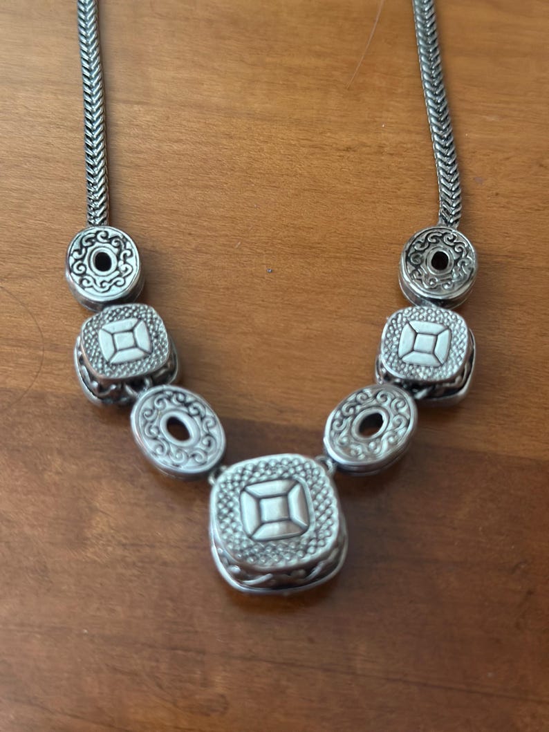 May include: Silver-toned necklace featuring a series of decorative elements. The necklace has a chain-like design with circular and square pendants. The pendants have intricate patterns and a central design. The necklace is displayed on a wooden surface.