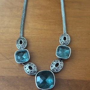 May include: Silver-toned necklace featuring a series of square, teal-colored gemstones. The necklace has a chain-like design with decorative circular accents between the gemstones. The gemstones are faceted, catching the light.