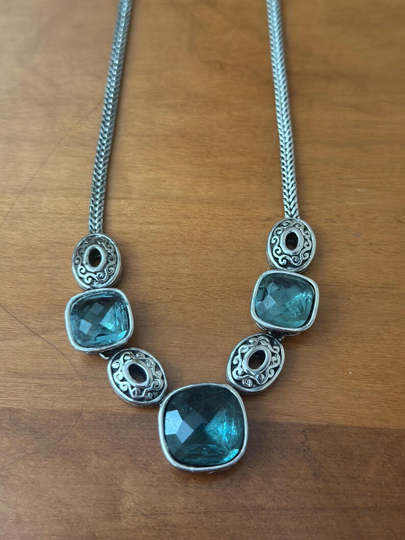 May include: Silver-toned necklace featuring a series of square, teal-colored gemstones. The necklace has a chain-like design with decorative circular accents between the gemstones. The gemstones are faceted, catching the light.