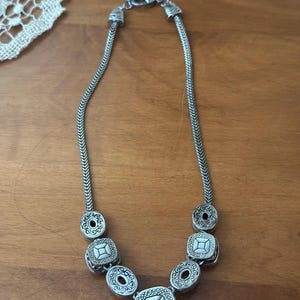 May include: Silver-toned necklace featuring a series of decorative elements. The necklace has a chain-like design with alternating round and square pendants. The pendants have intricate patterns and a vintage aesthetic. The necklace is displayed on a wooden surface.