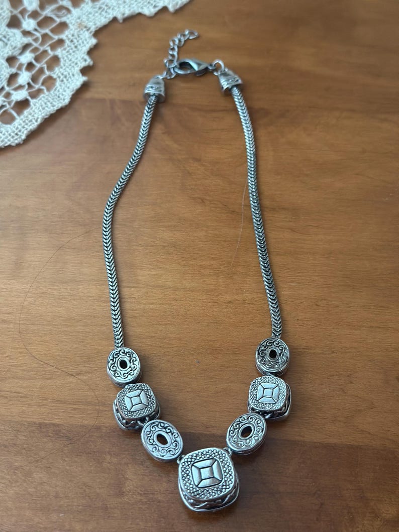 May include: Silver-toned necklace featuring a series of decorative elements. The necklace has a chain-like design with alternating round and square pendants. The pendants have intricate patterns and a vintage aesthetic. The necklace is displayed on a wooden surface.
