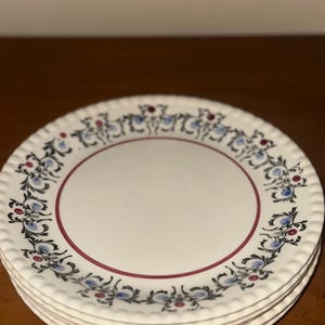 May include: A stack of white ceramic plates with a decorative floral pattern in blue, red, and black. The plates have a scalloped edge and a red inner border. The plates are stacked on top of each other.