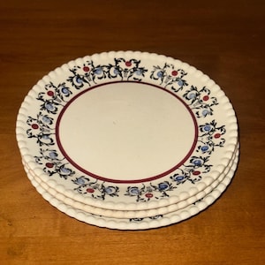 May include: Three white ceramic plates with a scalloped edge, stacked on a wooden surface. Each plate has a floral pattern in blue, red, and black around the rim, with a burgundy inner ring. The plates are stacked.